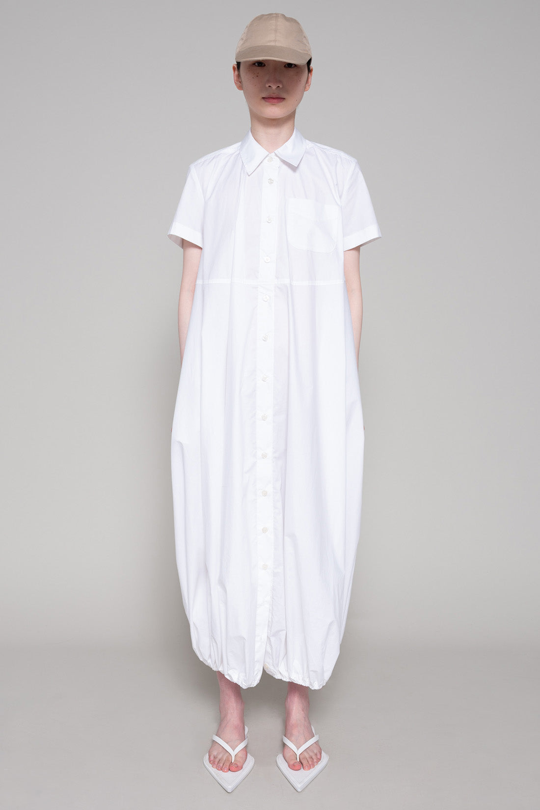 Person wearing a white dress with flip flops and beige cap on plain background