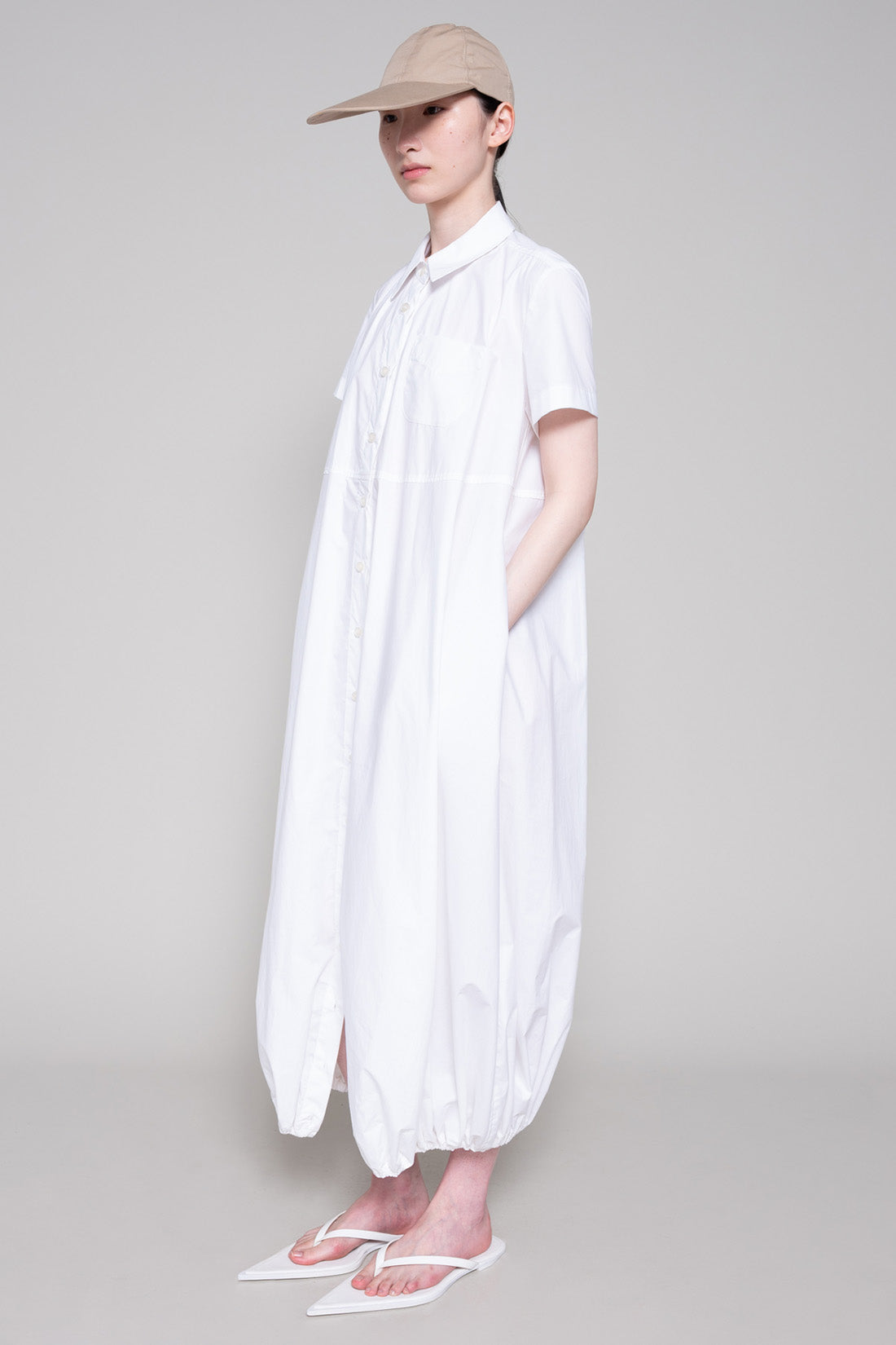 Person wearing a white dress with flip flops and beige cap on plain background