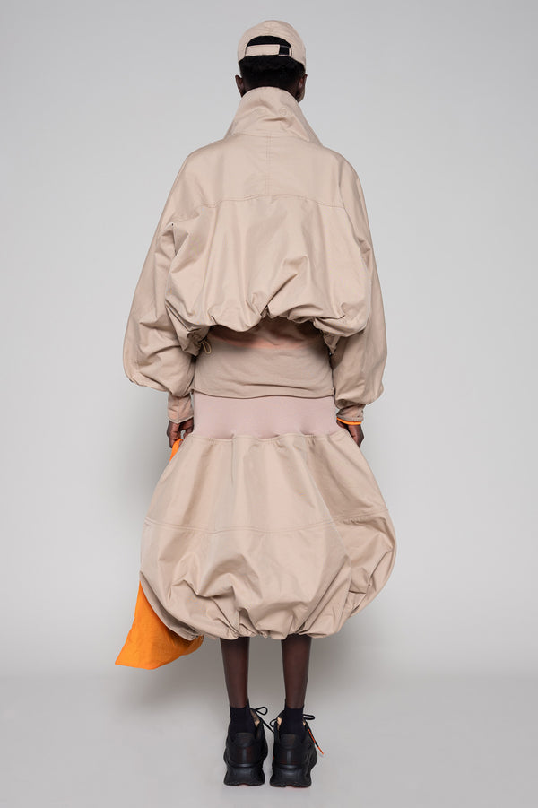 Person wearing a beige jacket, skirt and cap with an orange tote bag and black sneakers against a plain background