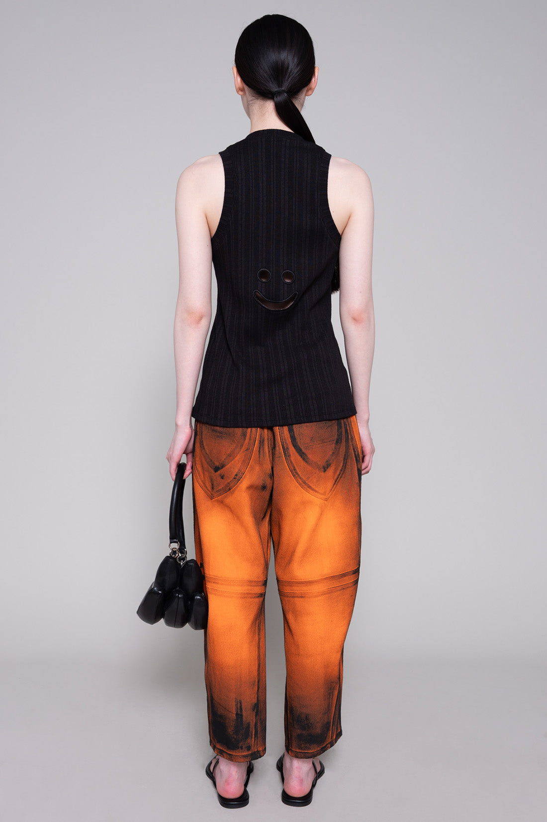 Person wearing a black sleeveless top and orange painted pants with black bag on a plain background