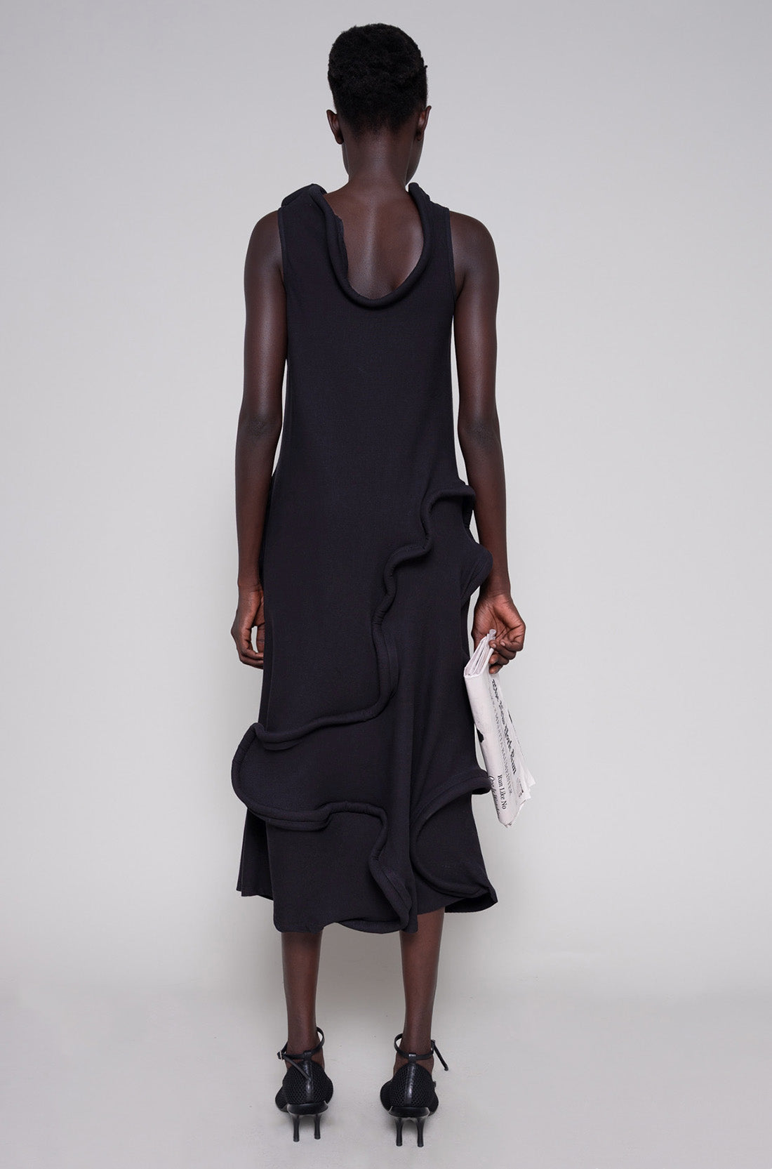Person wearing a black dress and heels holding a tote bag on a plain background