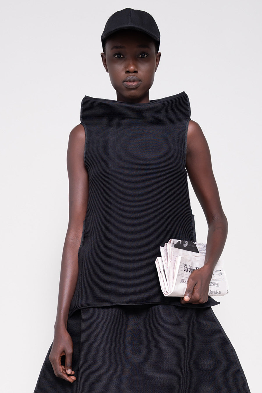 Person wearing a black sleeveless top and skirt with  cap holding a newspaper against a plain background