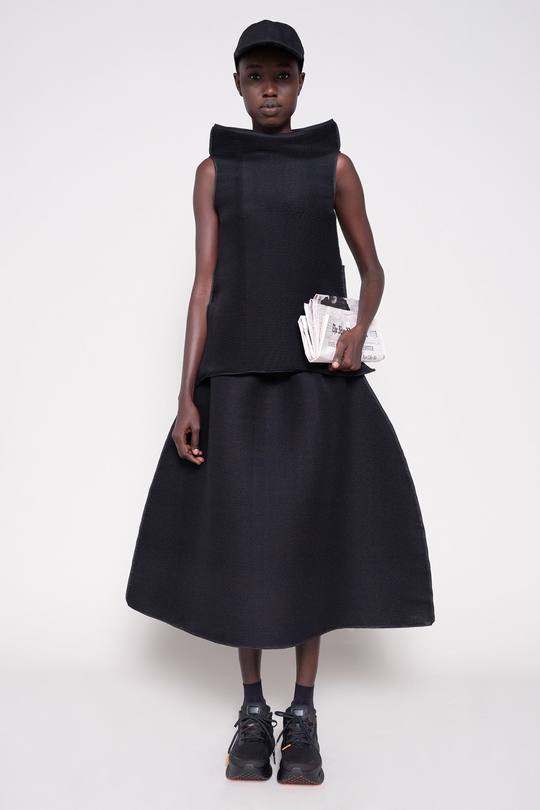 Person wearing a black sleeveless top and skirt with  cap holding a newspaper against a plain background