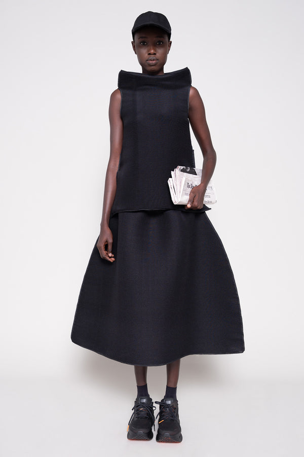Person wearing a black sleeveless top and skirt with  cap holding a newspaper against a plain background