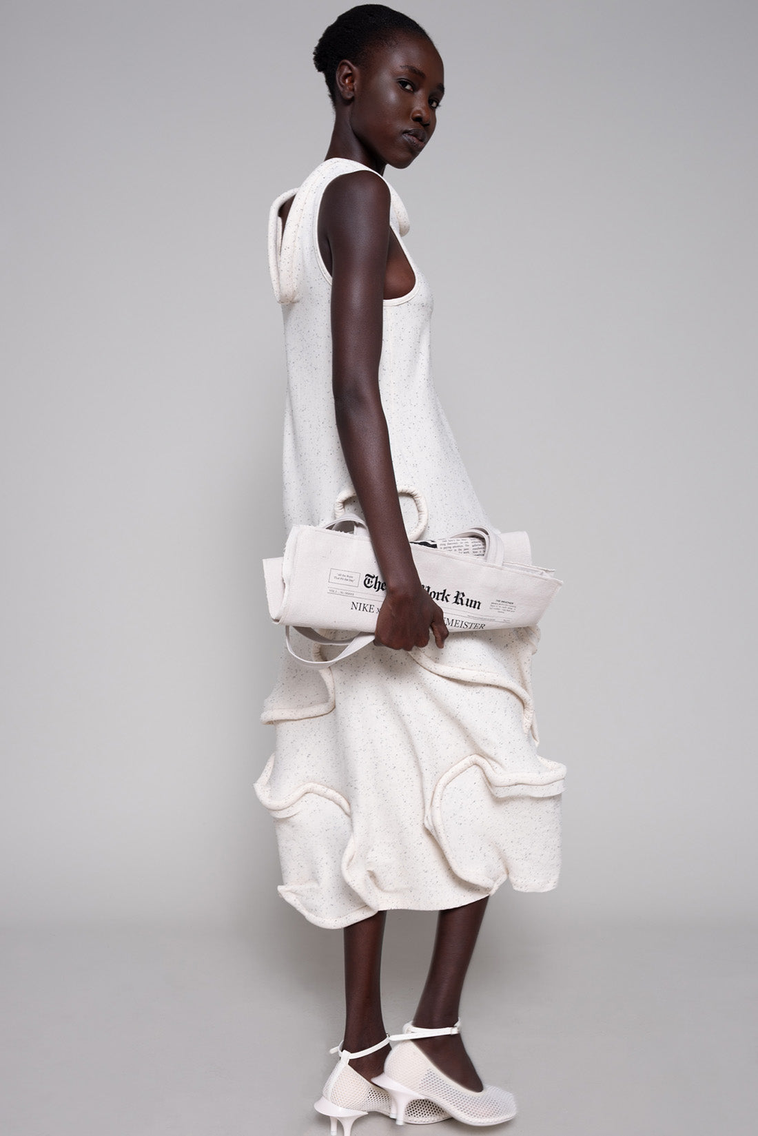 Person wearing a white dress and heels holding a tote bag on a plain background