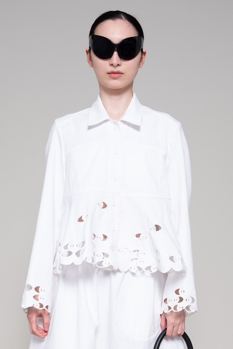 Person with black sunglasses wearing a white shirt and skirt with cut-out details holding a black bag on a plain background