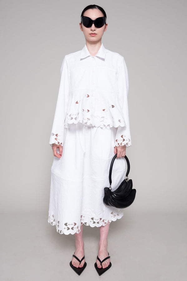 Person with black sunglasses wearing a white shirt and skirt with cut-out details holding a black bag on a plain background