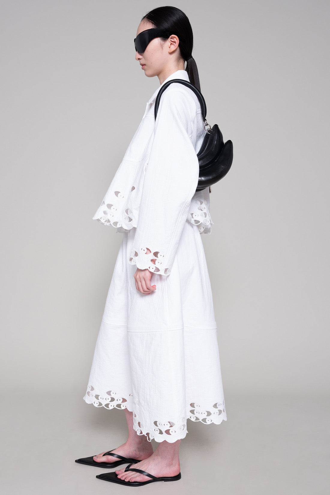 Person with black sunglasses wearing a white shirt and skirt with cut-out details holding a black bag on a plain background