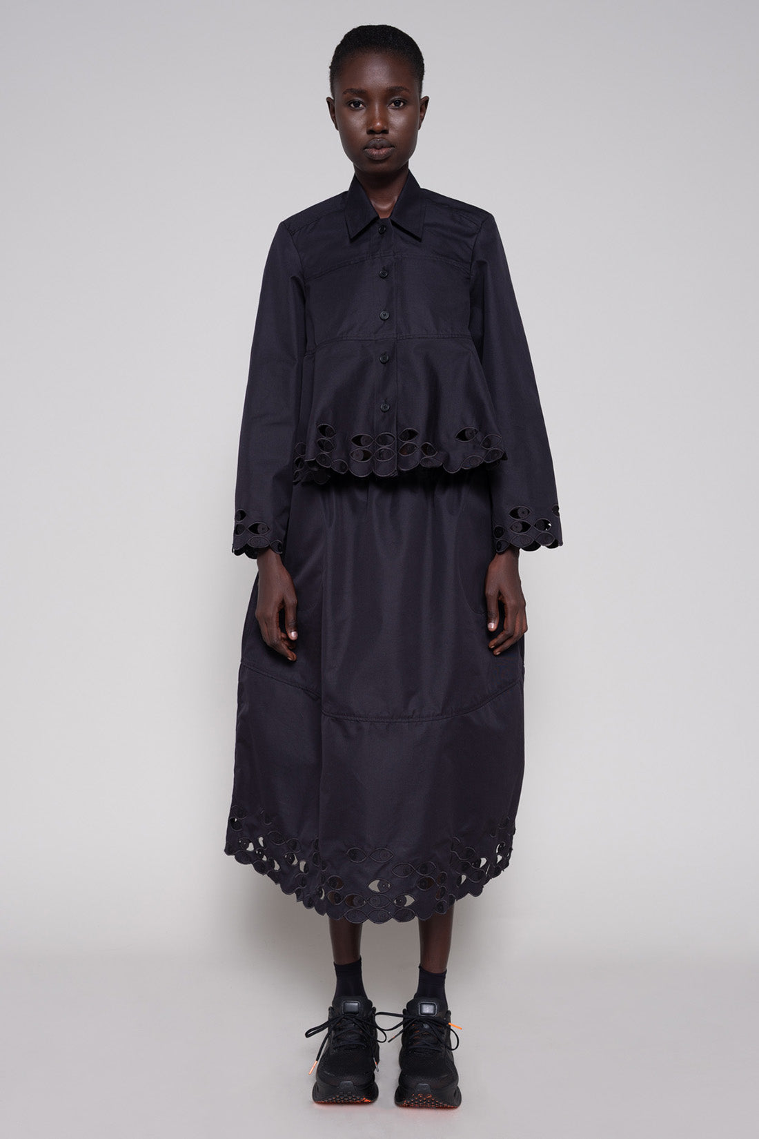 Person wearing a black shirt and skirt with cut-out details on a plain background