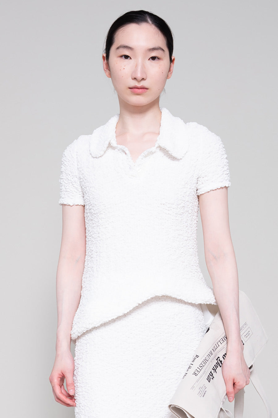 Person wearing a textured white polo and skirt, holding a tote bag on a plain background.
