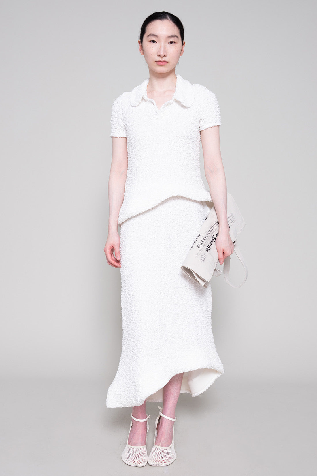 Person wearing a textured white polo and skirt with heels, holding a tote bag on a plain background.