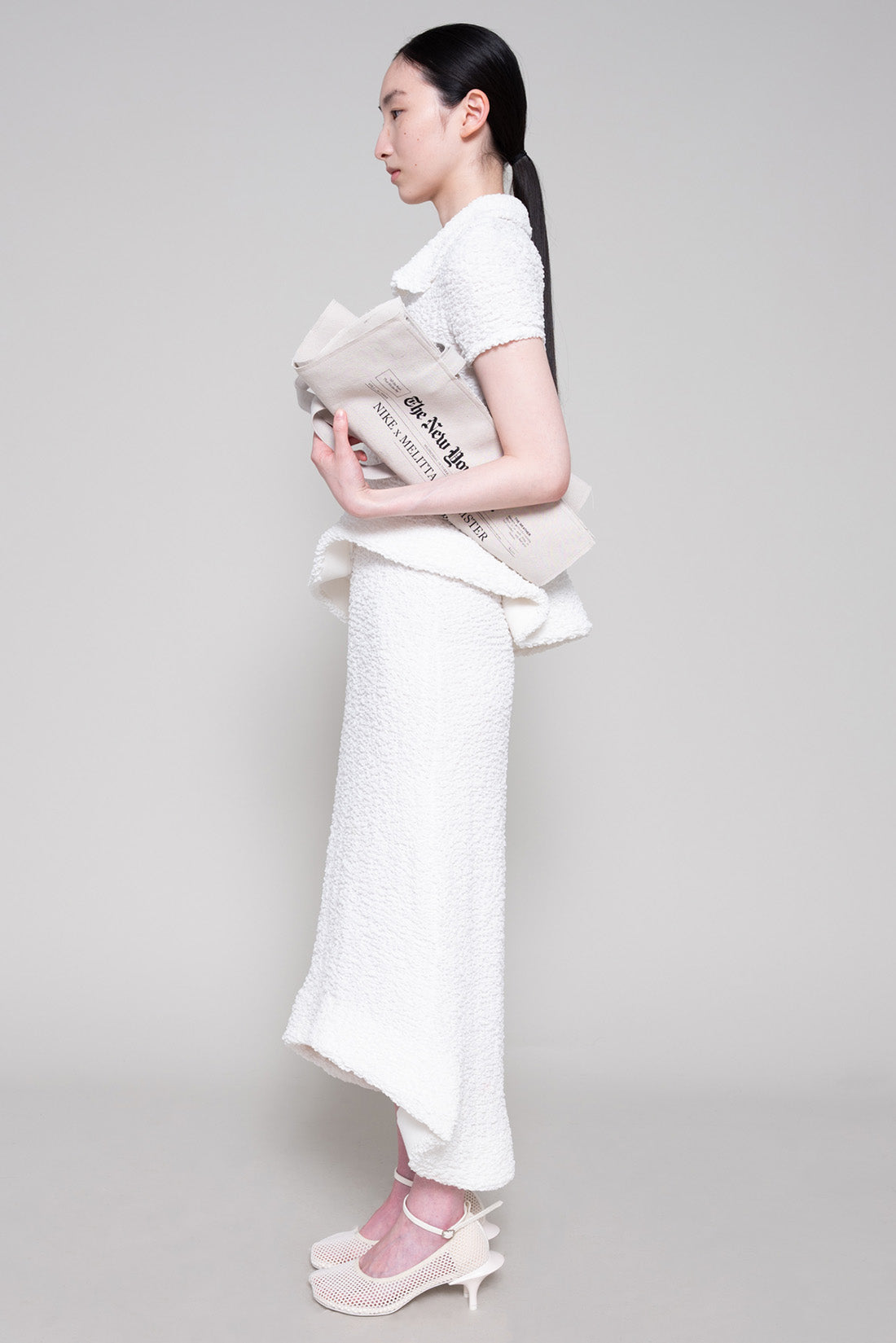 Person wearing a textured white polo and skirt with heels, holding a tote bag on a plain background.