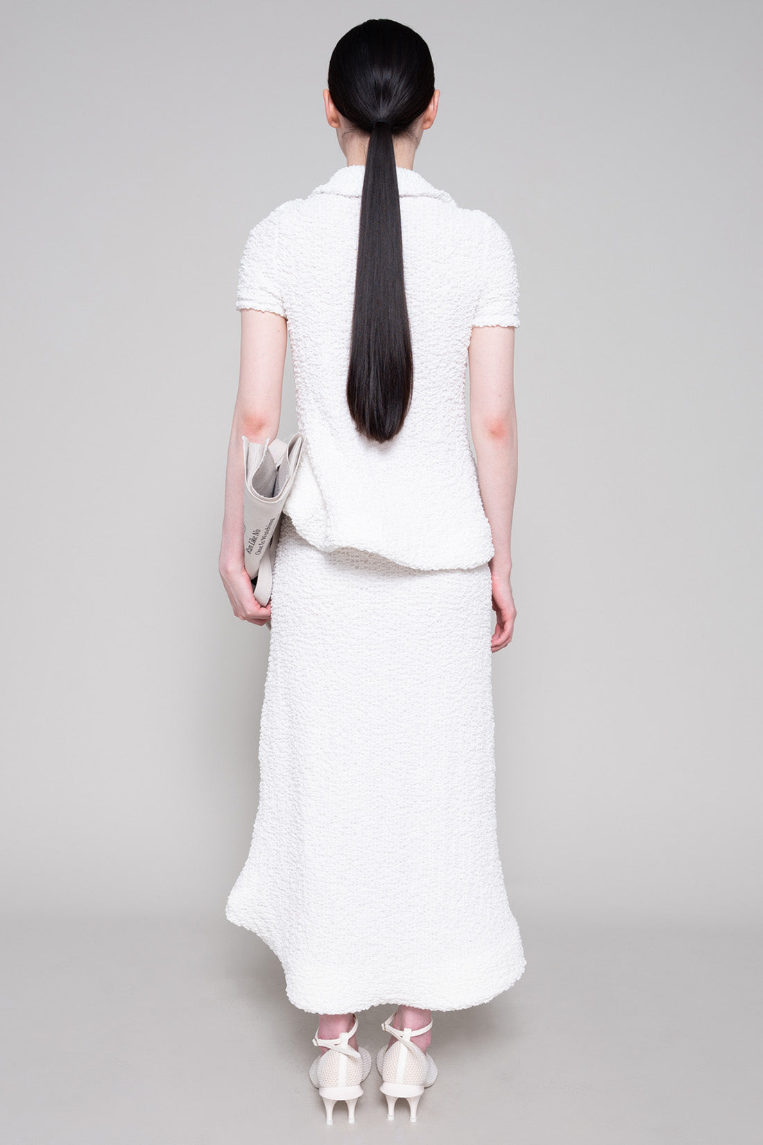 Person wearing a textured white polo and skirt with heels, holding a tote bag on a plain background.