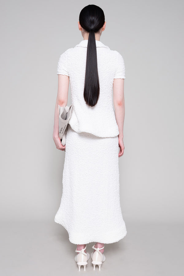 Person wearing a textured white polo and skirt with heels, holding a tote bag on a plain background.