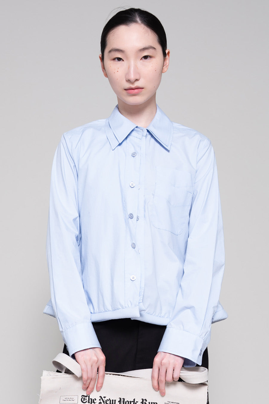Person wearing a light blue shirt and black pants holding a tote with newspaper print against a plain background