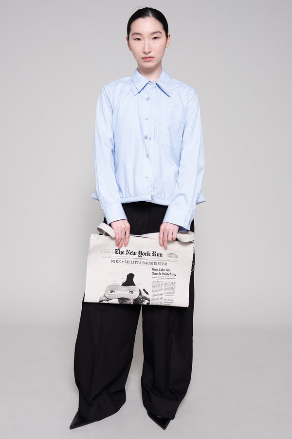 Person wearing a light blue shirt and black pants holding a tote with newspaper print against a plain background