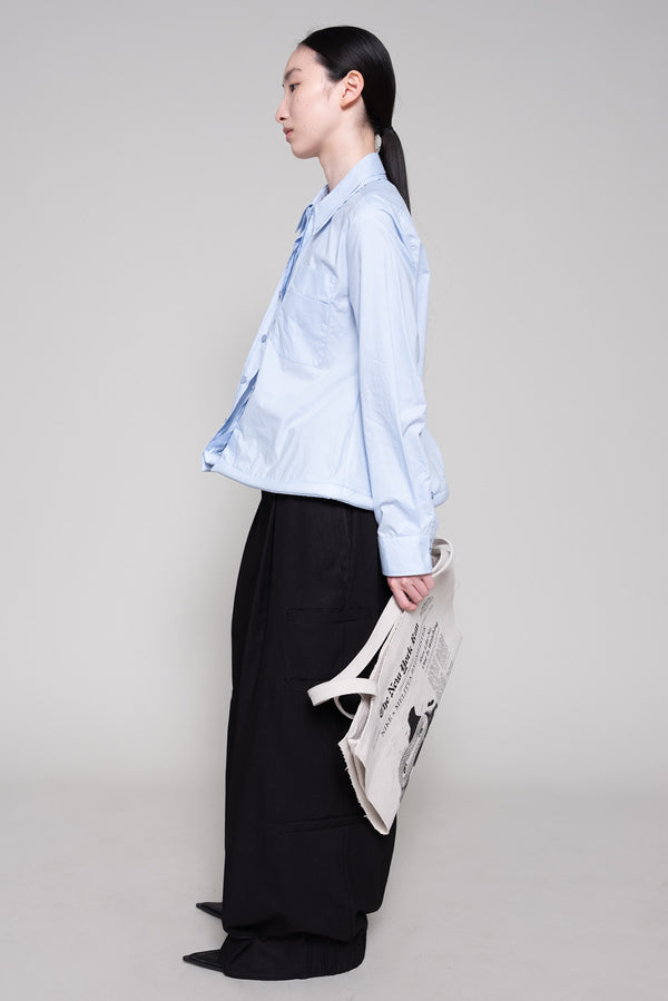 Person wearing a light blue shirt and black pants holding a tote with newspaper print against a plain background