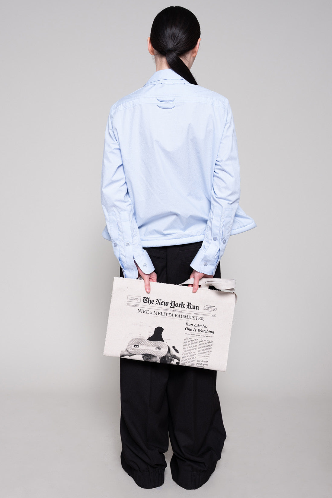 Person wearing a light blue shirt and black pants, holding a tote bag with newspaper print on a plain background