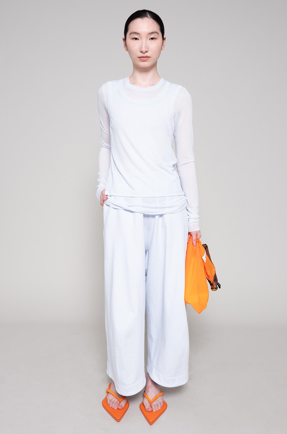 Person wearing a light long sleeve and sweatpants with orange flip flops and a tote bag on a plain background