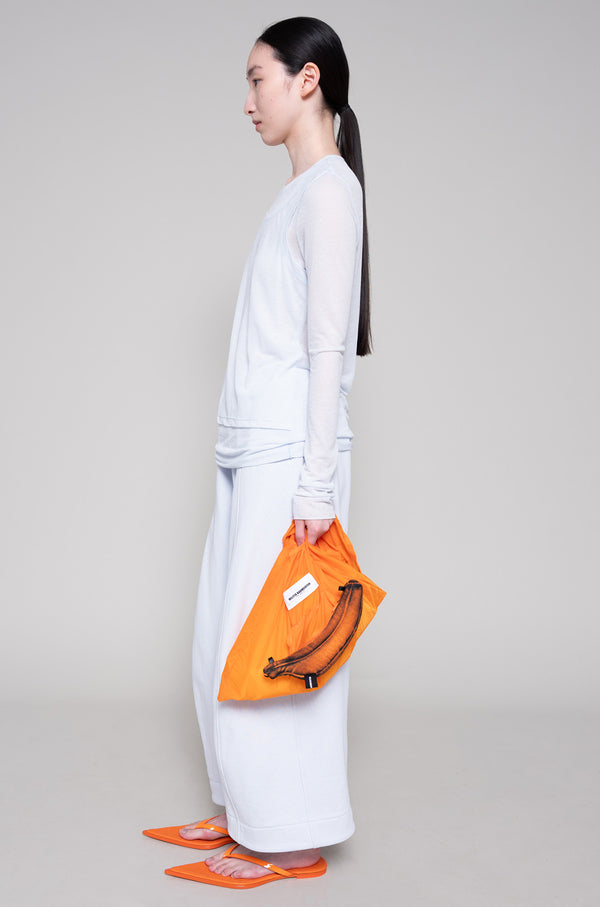 Person wearing a light longsleeve and sweatpants with orange flip flops,holding an orange bag against a plain background