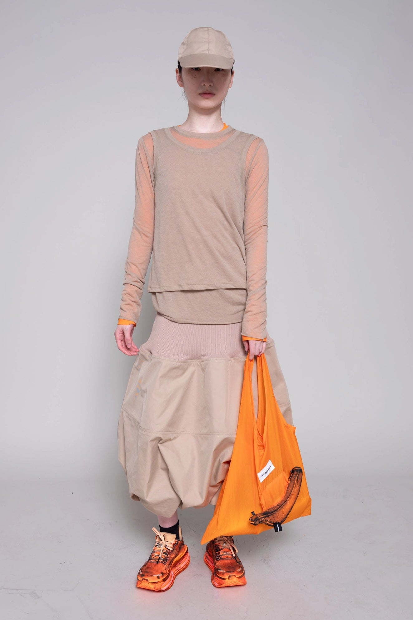 Person wearing a beige outfit with orange accents, holding an orange bag on a gray background