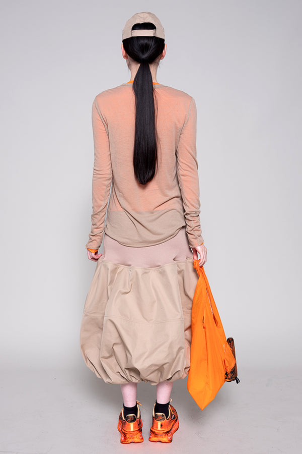 Person wearing a beige outfit with a ponytail, holding an orange bag on a plain background