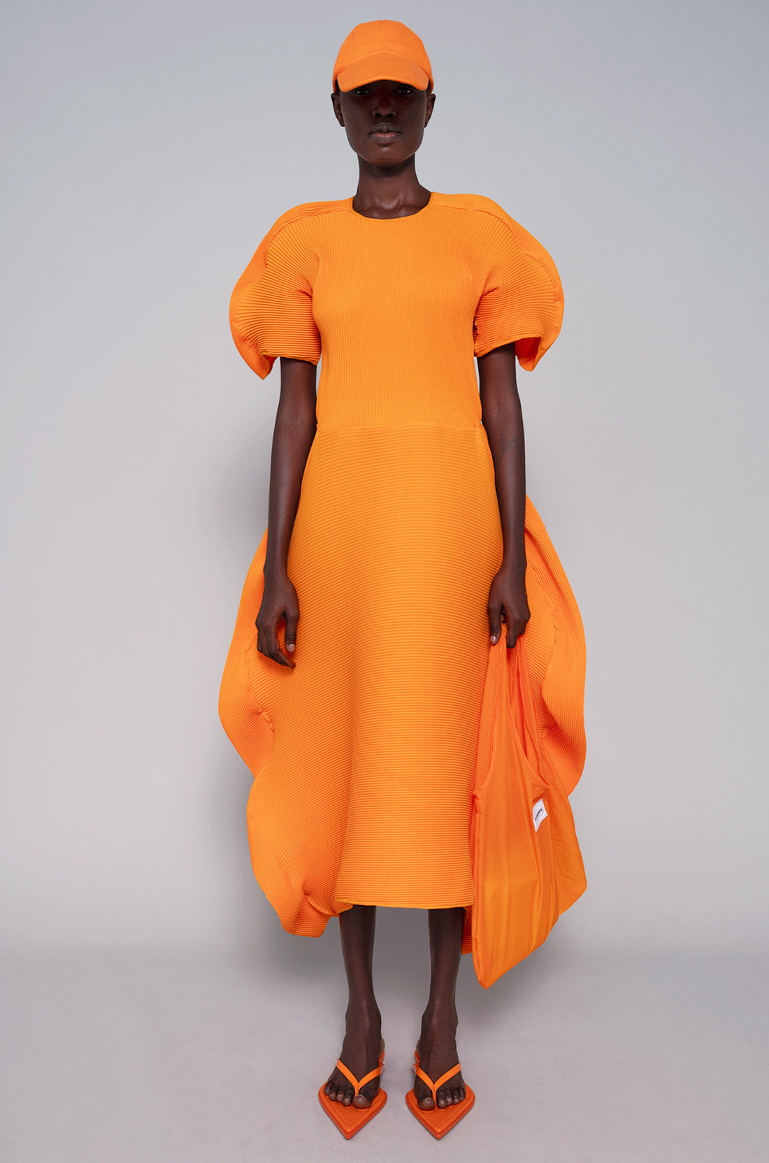 Person wearing an orange dress, tote bag, flip flops and cap on a gray background