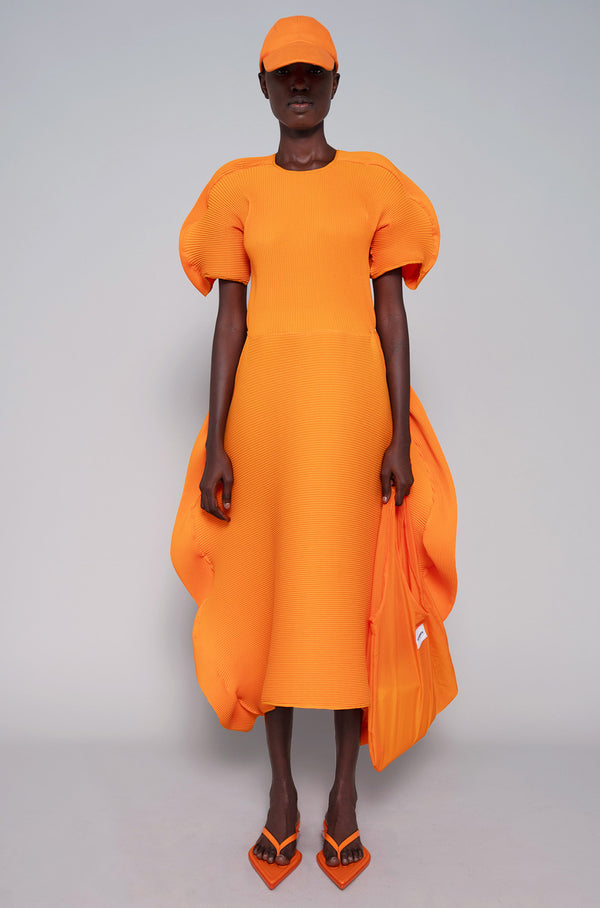 Person wearing an orange dress, tote bag, flip flops and cap on a gray background