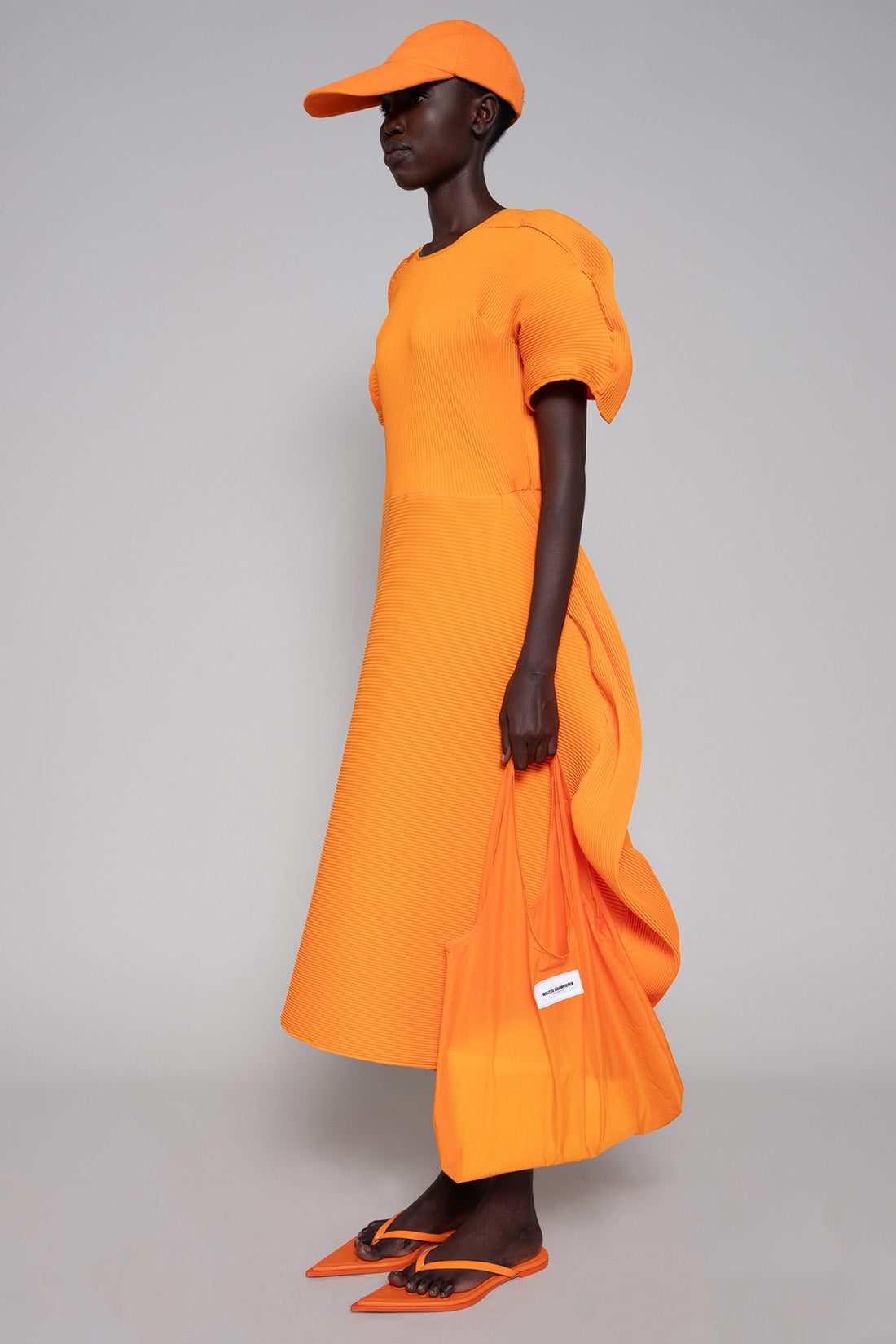 Person wearing an orange dress, tote bag, flip flops and cap on a gray background