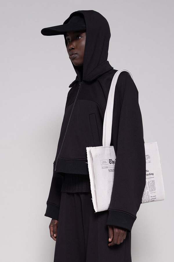 Person wearing a black hoodie and cap, holding a white tote bag with text, on a light gray background