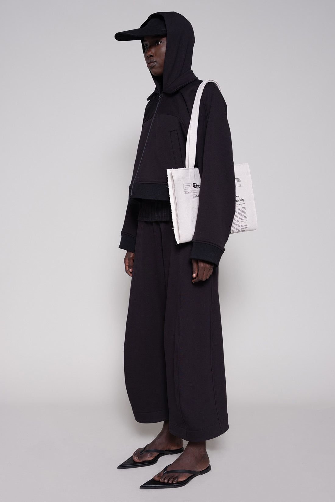 Person wearing a black hoodie, jacket and sweatpants with a cap and tote against a plain background