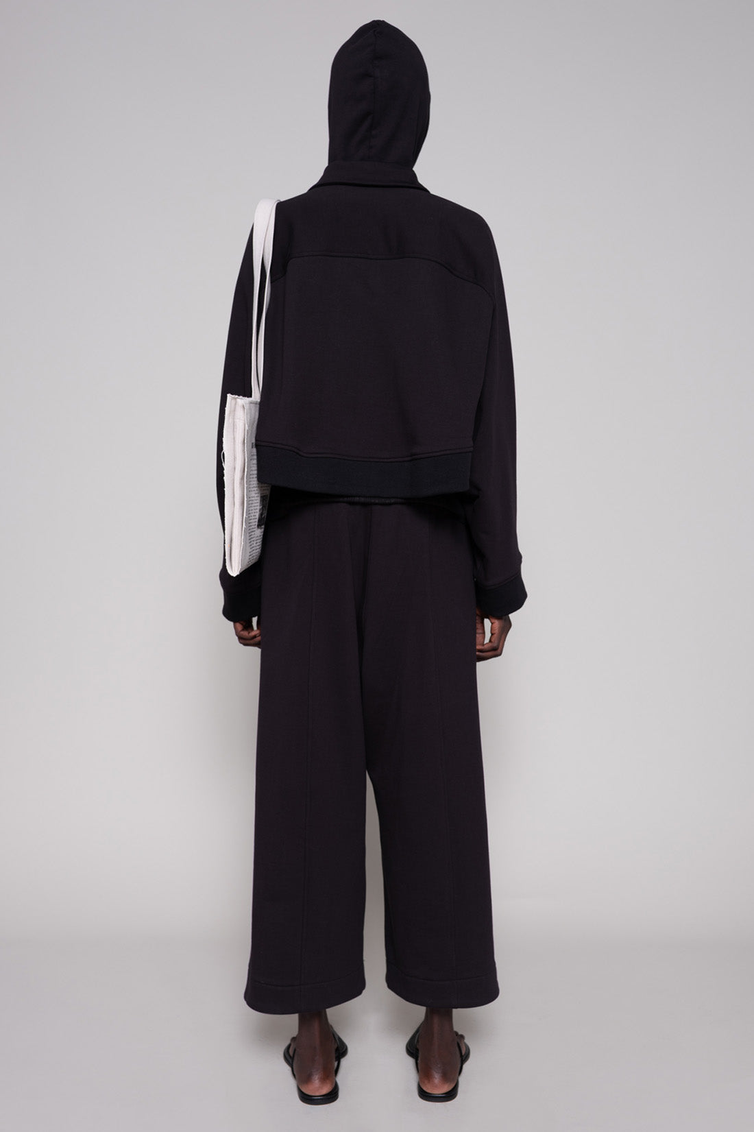 Person wearing a black hoodie, jacket and sweatpants with a tote against a plain background