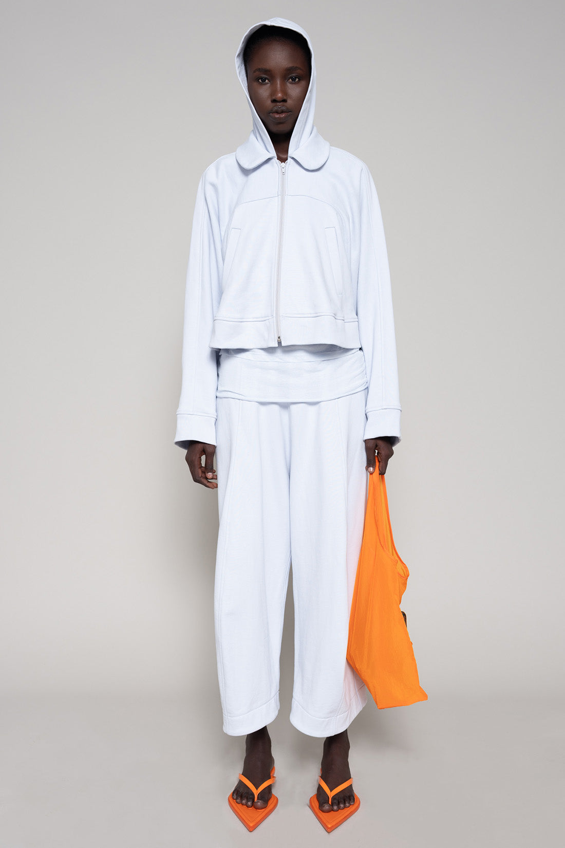 Person wearing a light blue hoodie and sweatpants  with orange shoes and an orange bag on a plain background