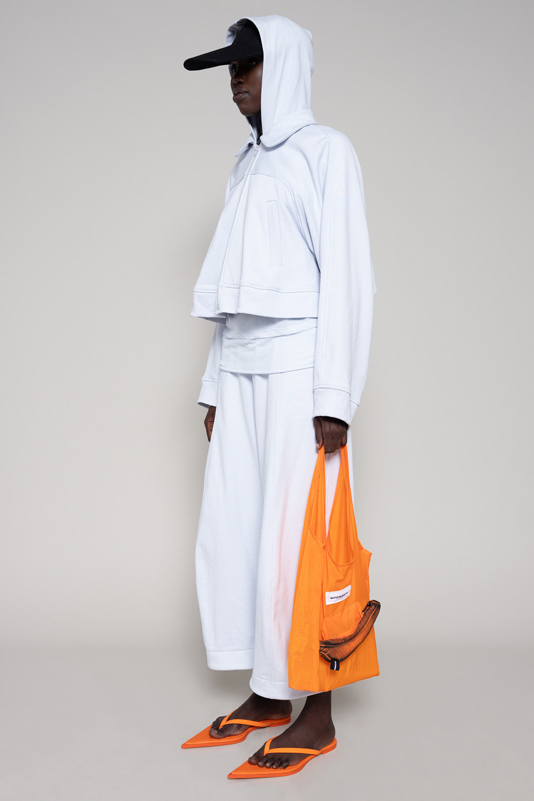 Person wearing a light blue hoodie and sweatpants  with a black cap, orange shoes and an orange bag on a plain background