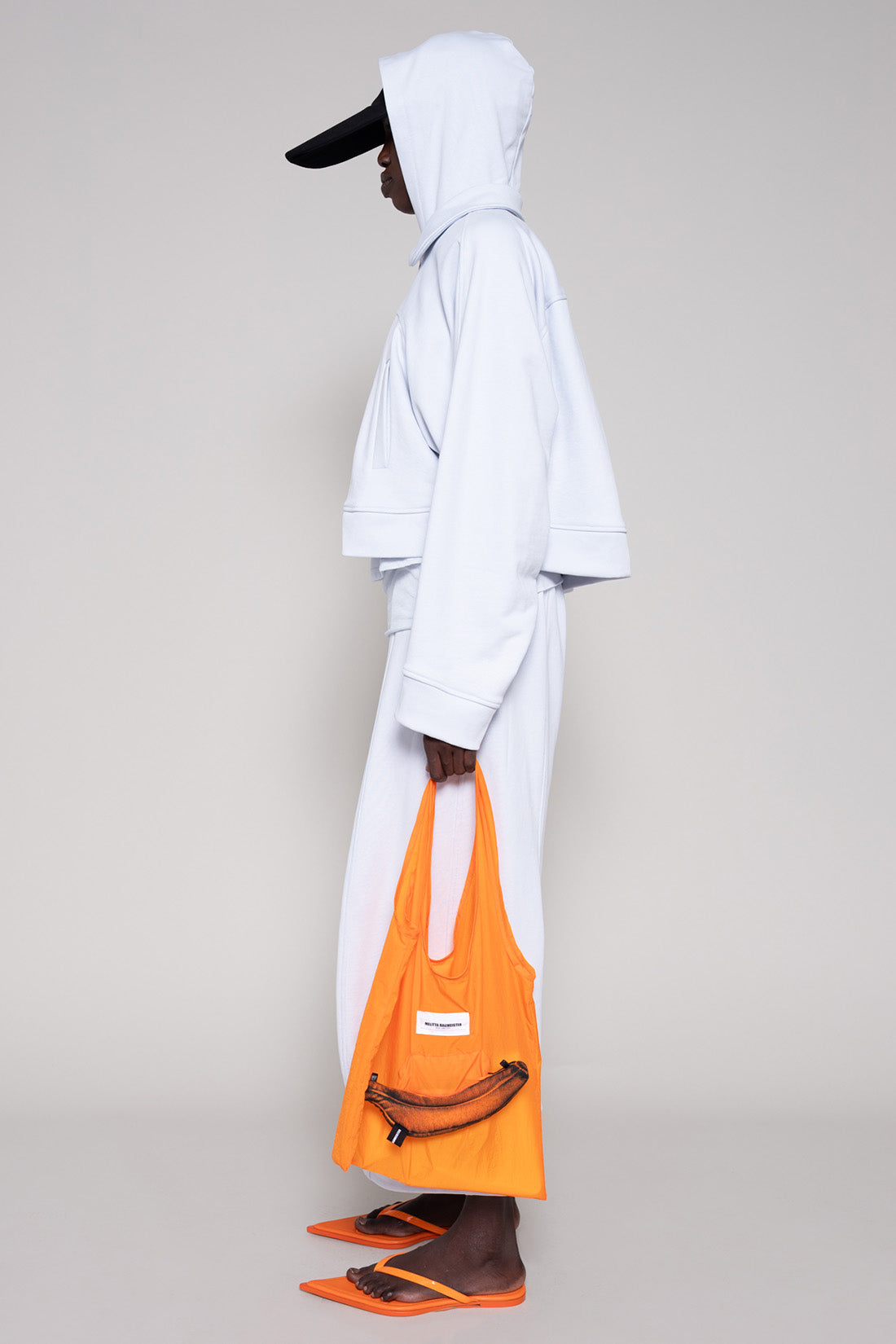 Person wearing a light blue hoodie and sweatpants  with a black cap, orange shoes and an orange bag on a plain background