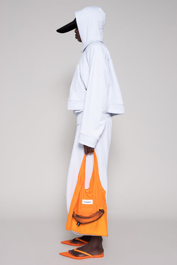Person wearing a light blue hoodie and sweatpants  with a black cap, orange shoes and an orange bag on a plain background
