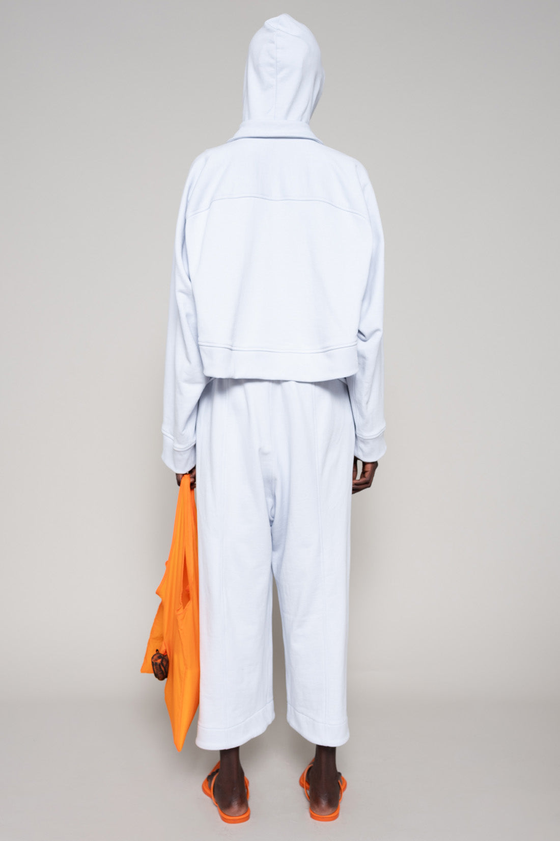Person wearing a light blue hoodie and sweatpants  with orange shoes and an orange bag on a plain background