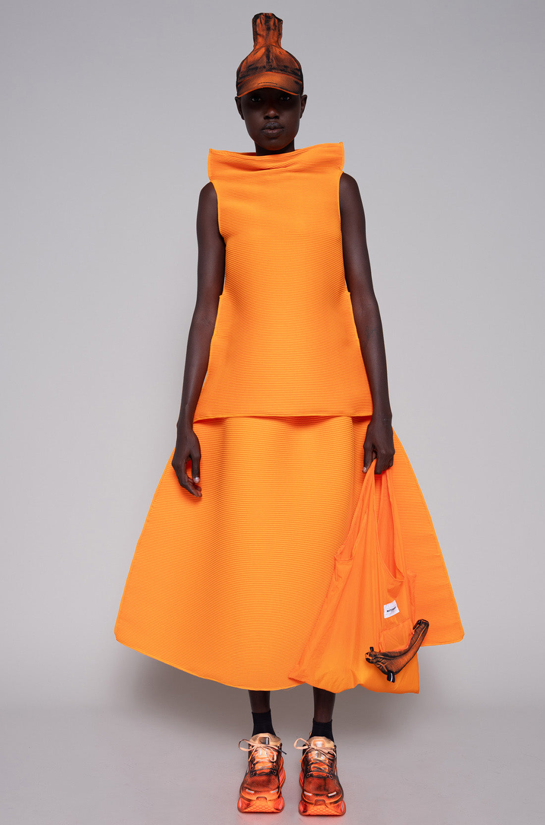 Person wearing a orange sleeveless top and skirt with painted hat, tote bag and shoes against plain background