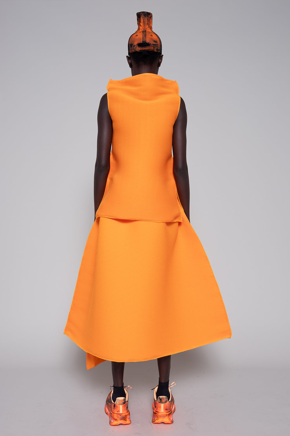 Person wearing an orange sleeveless dress with a hood on a gray background