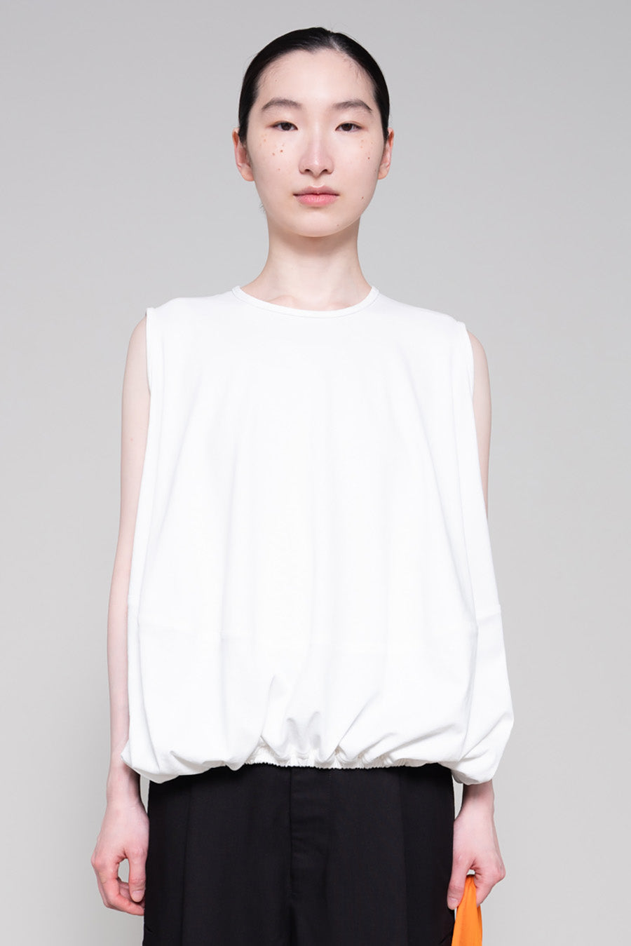 Person wearing a white sleeveless top and black pants, holding an orange bag on a gray background