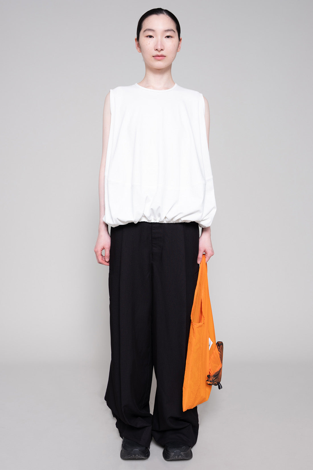 Person wearing a white sleeveless top and black pants, holding an orange bag on a gray background