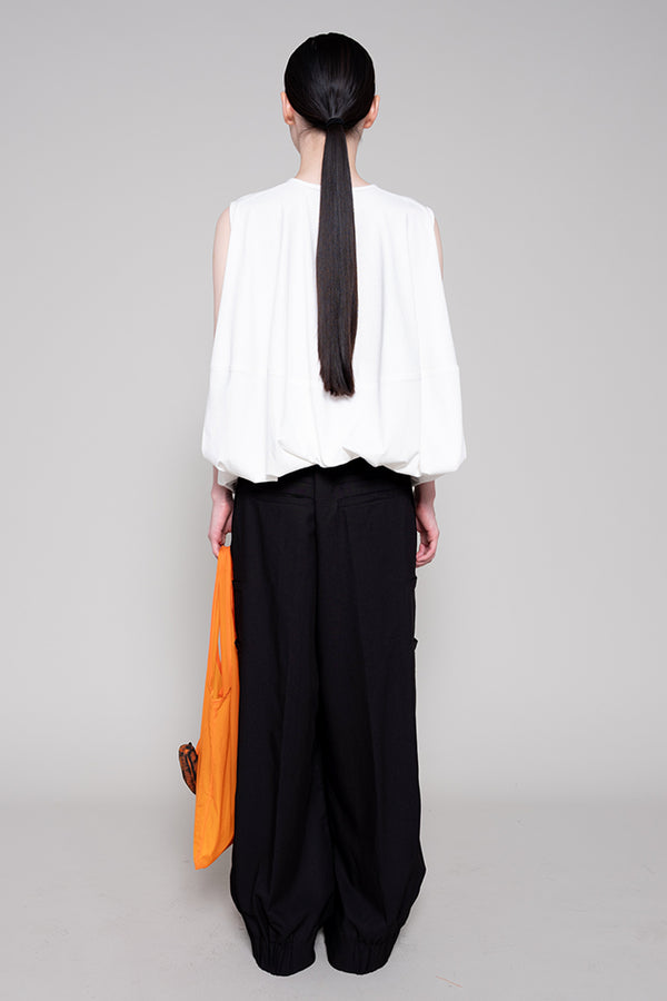 Person wearing a white sleeveless top and black pants with an orange section, standing against a gray background.