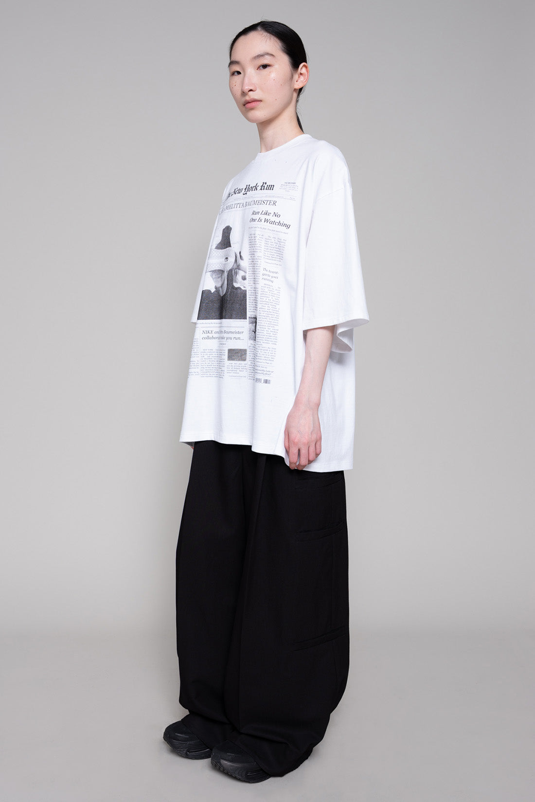 Person wearing a white oversized shirt with black text and graphics, paired with black pants on a gray background