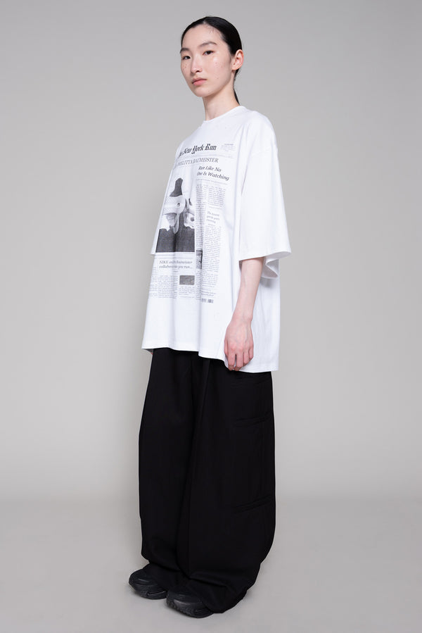 Person wearing a white oversized shirt with black text and graphics, paired with black pants on a gray background