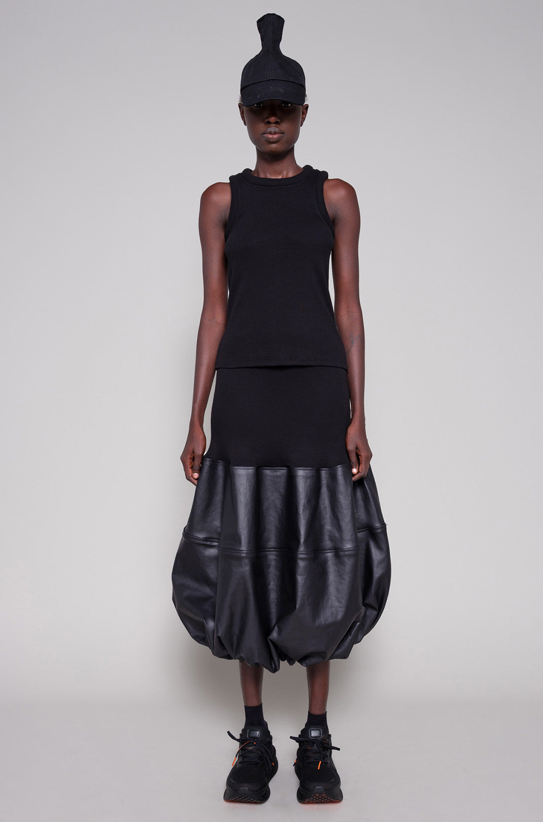 Person wearing a black sleeveless top and black leather skirt with a cap on a plain background