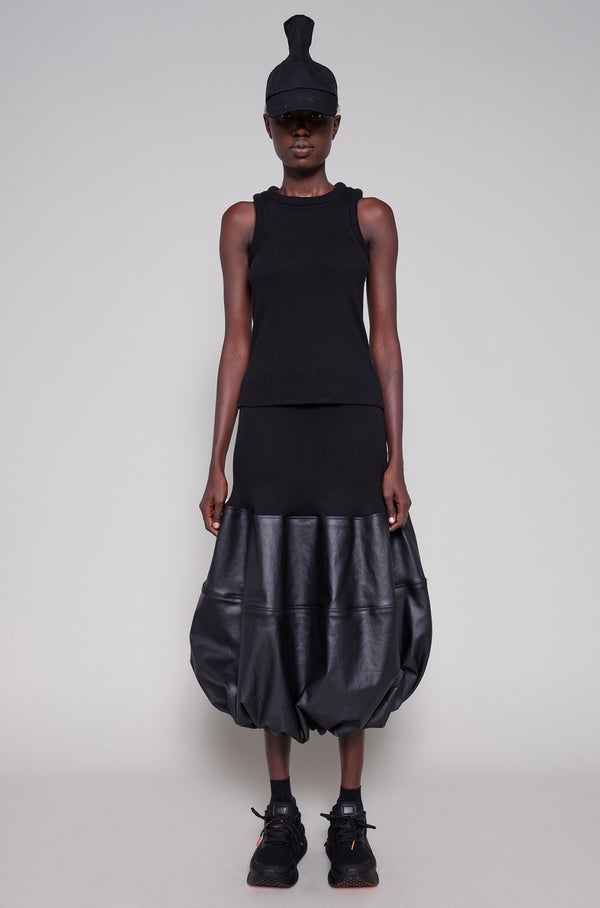 Person wearing a black sleeveless top and black leather skirt with a cap on a plain background