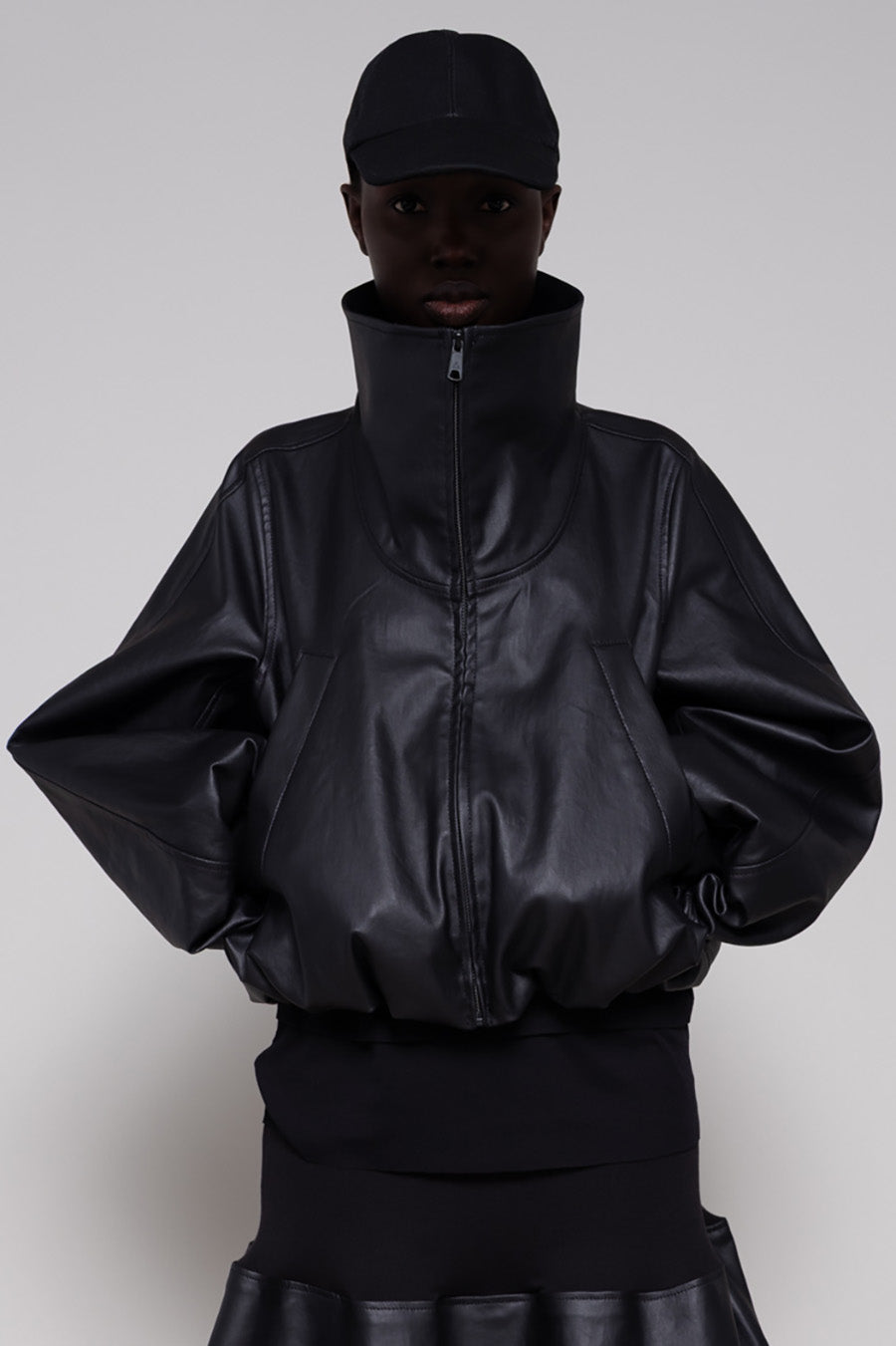 Person wearing a black leather jacket and skirt with a cap on a plain background