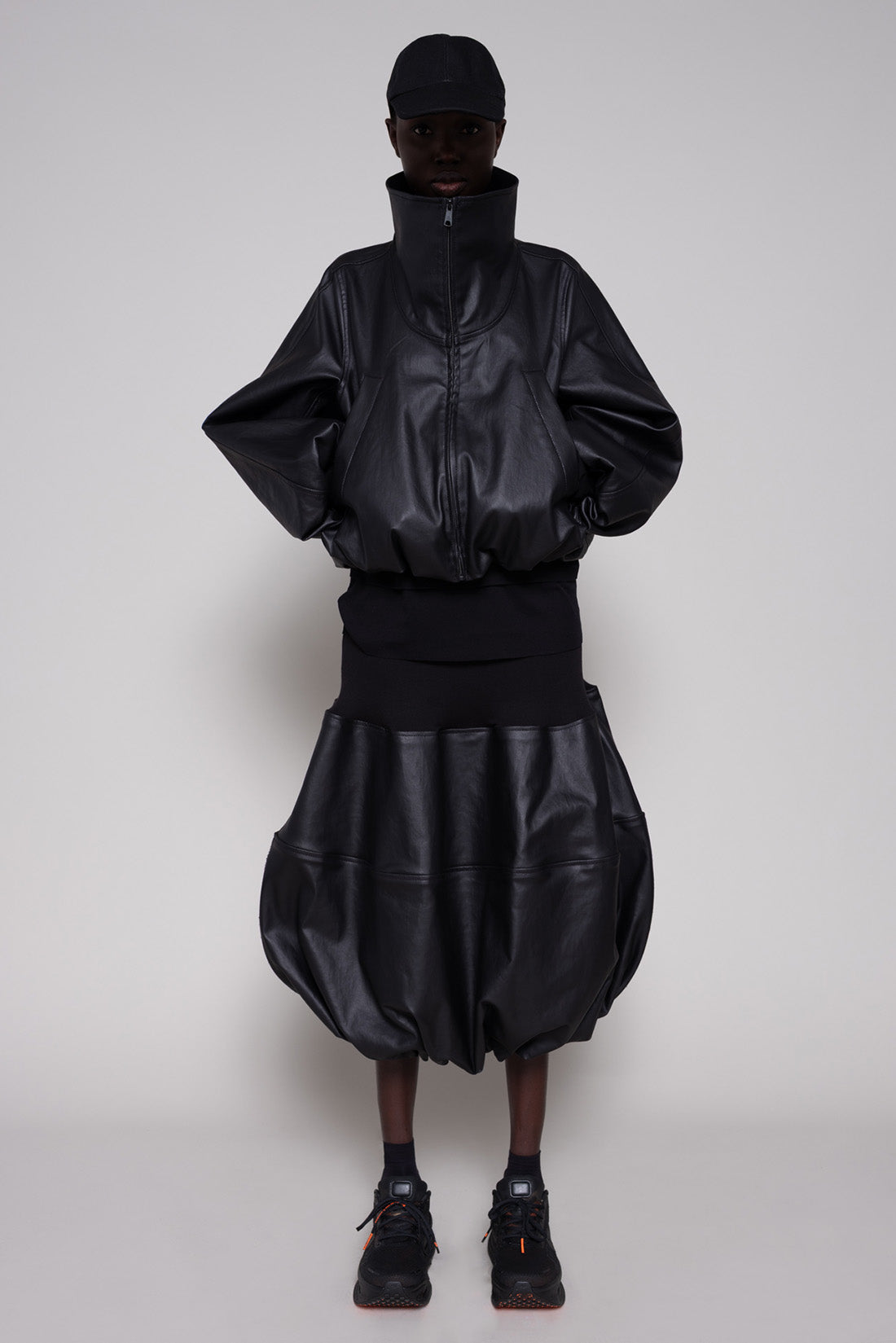 Person wearing a black leather jacket and skirt with a cap on a plain background