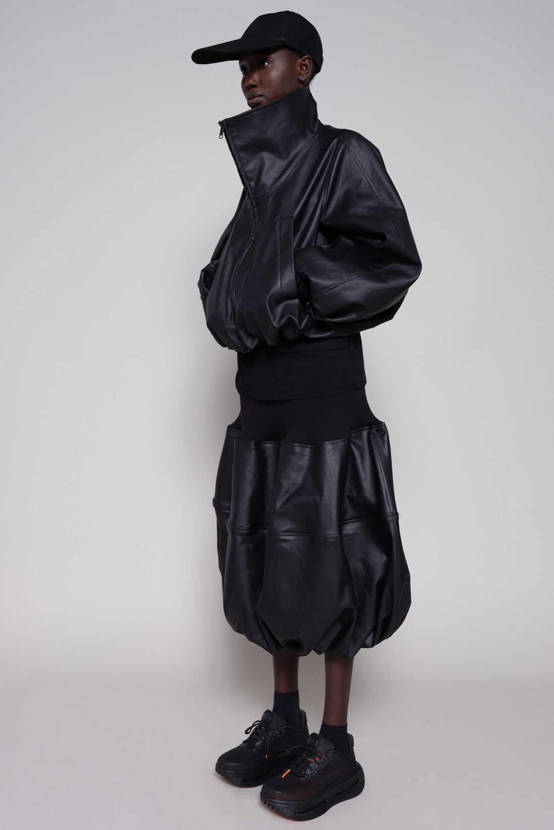 Person wearing a black leather jacket and skirt with a cap on a plain background