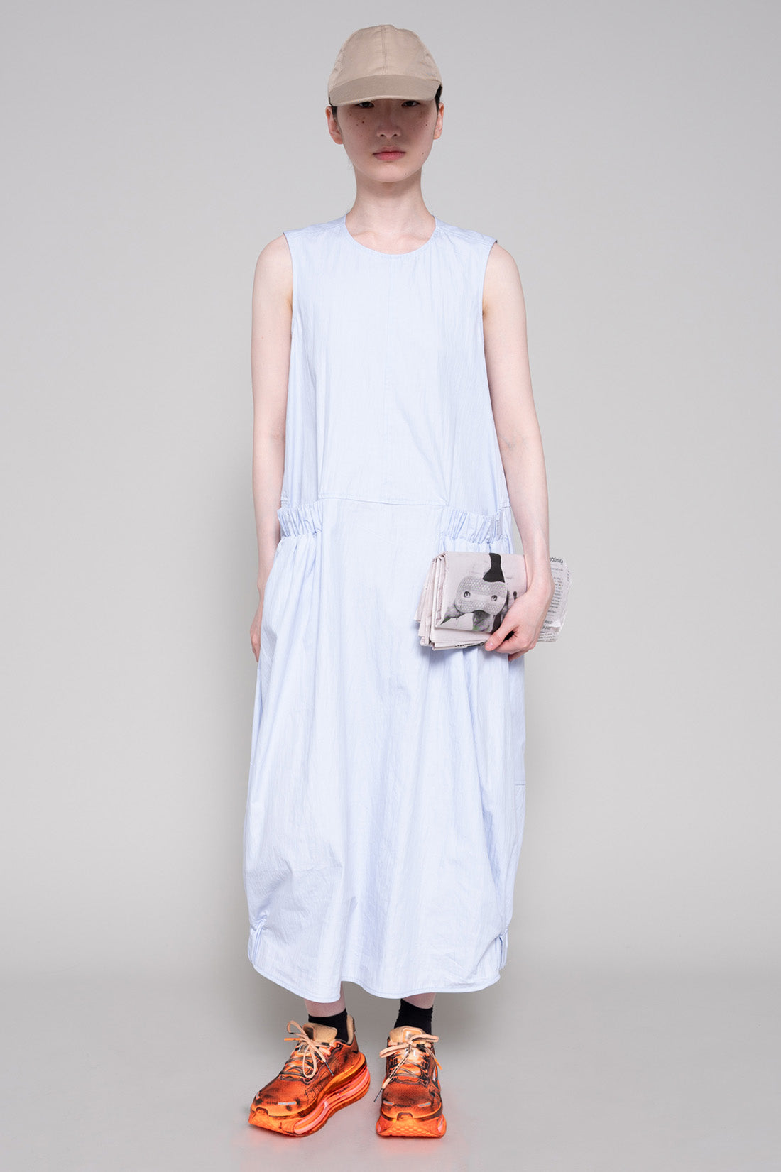 Person wearing a light blue sleeveless dress with beige cap and orange sneakers, holding a newspaper against a plain background.